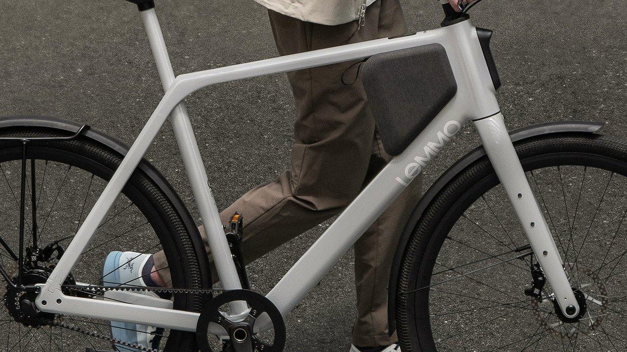 A person standing beside a gray Lemmo electric bicycle with black fenders and a frame-mounted battery.