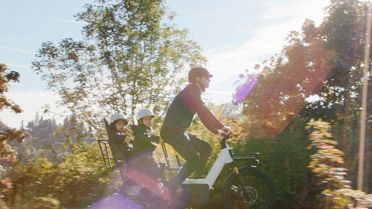 An adult rides an electric cargo bike carrying two helmeted children through a sunny, tree-lined area.