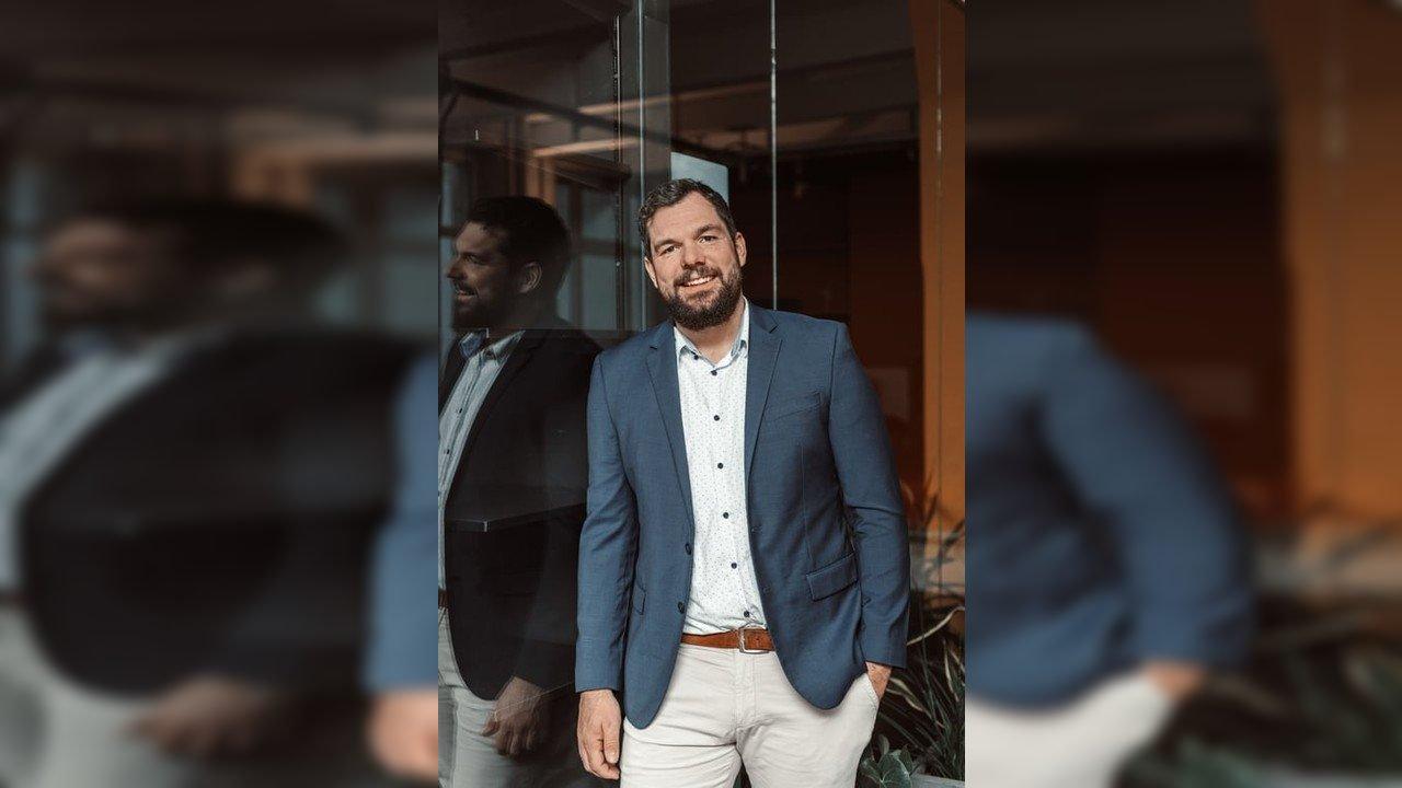 Jan Steggewentz in a blue blazer standing beside a glass wall with his reflection visible.