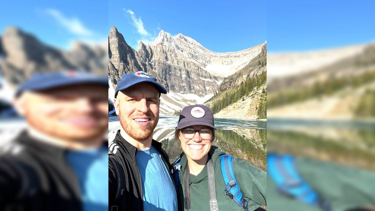Two hikers wearing caps and backpacks stand before a partly frozen mountain lake and rocky peaks.