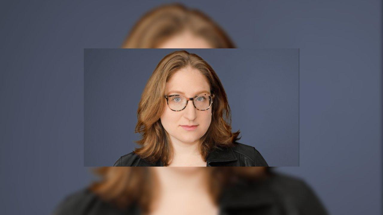 Rebecca Brocato wearing glasses and a black jacket against a blue-gray background.