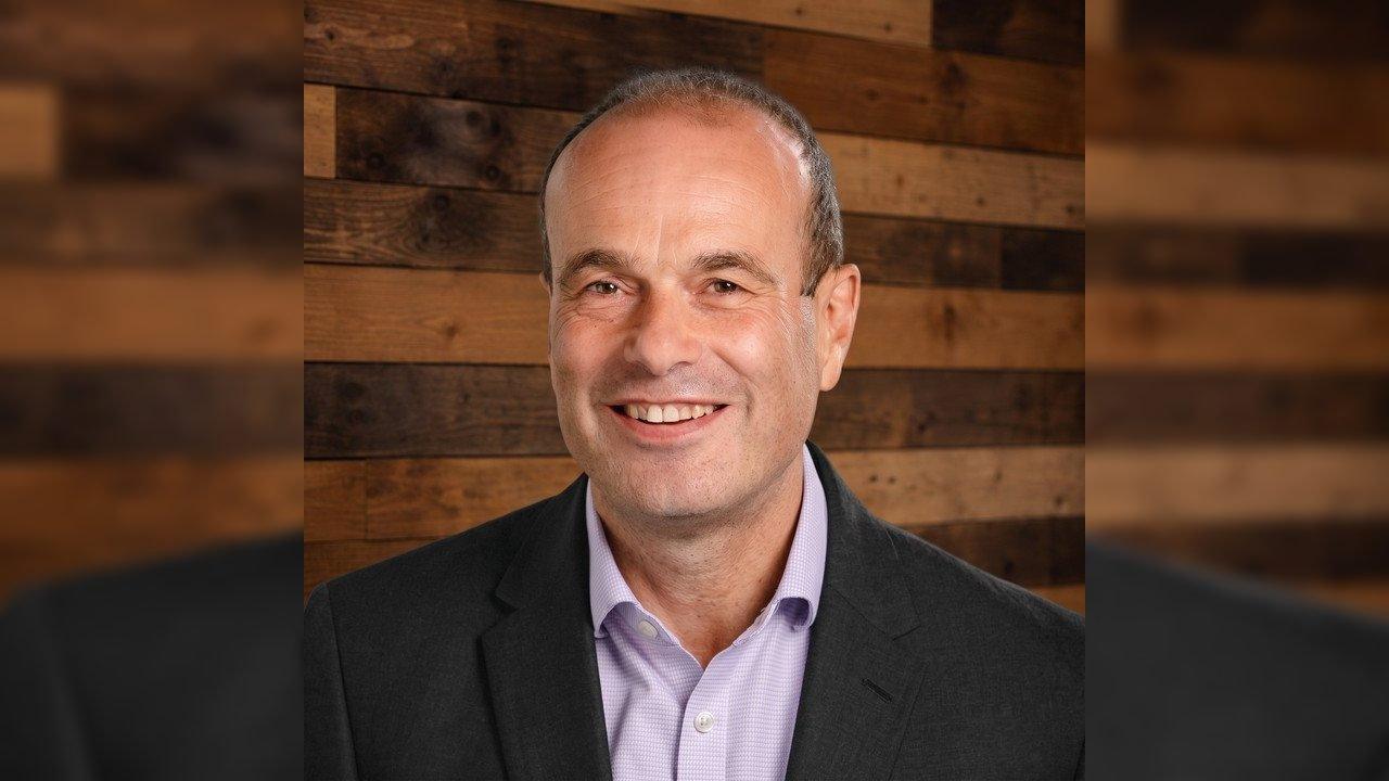 Smiling man in a dark jacket and light shirt against a wooden plank wall
