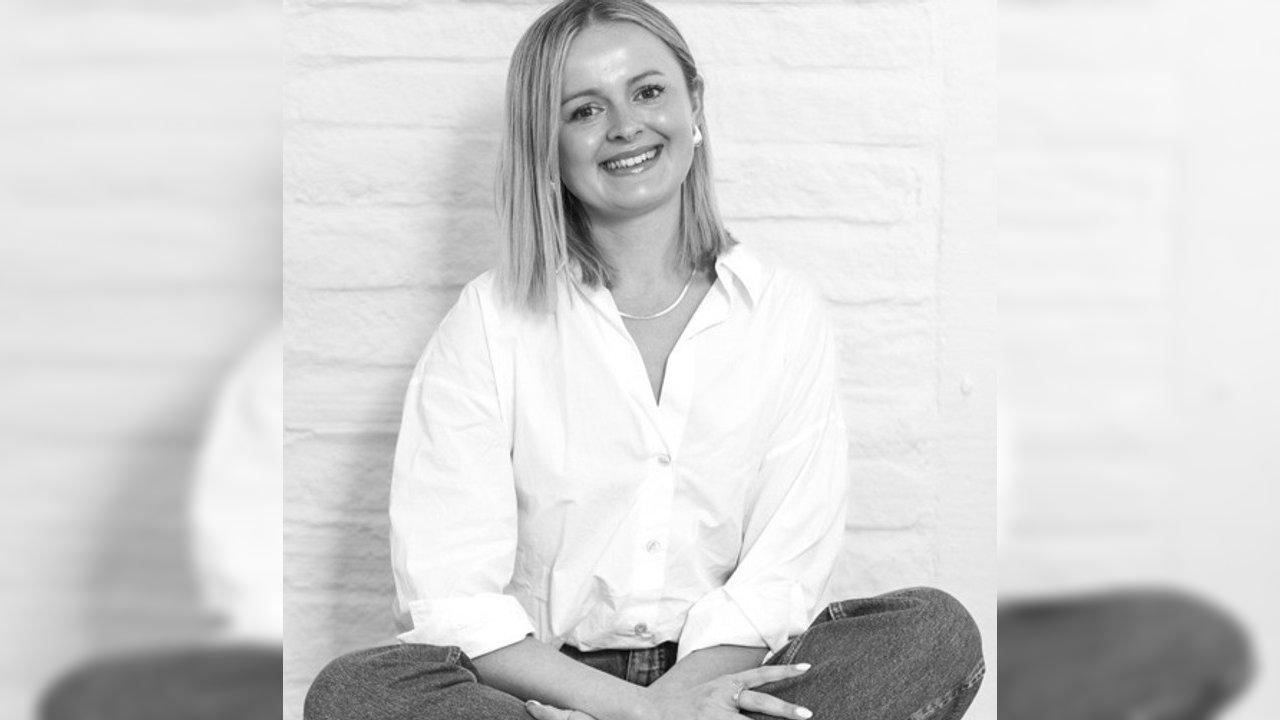 Smiling woman in a white button-down shirt and jeans seated against a white brick wall