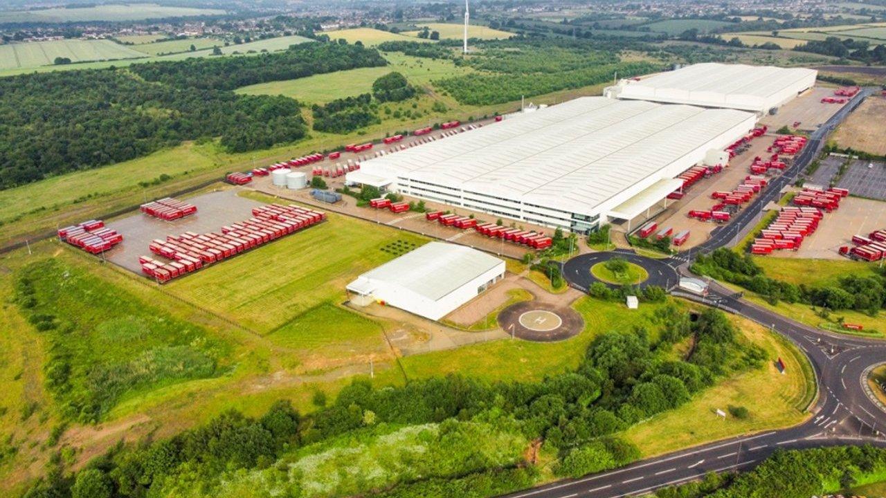 A large warehouse surrounded by rows of red trucks, fields and woodland, with a helipad at the entrance.