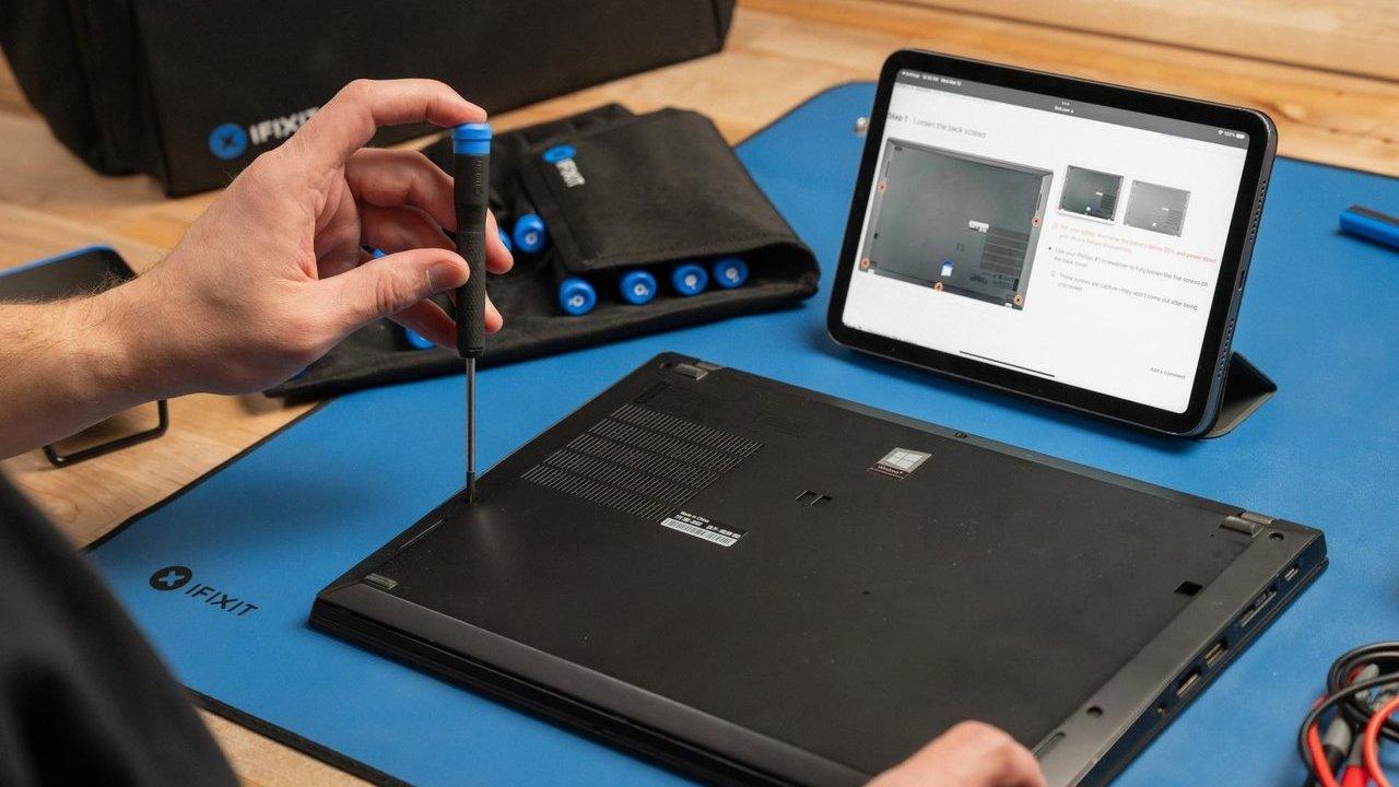 Person removing a laptop bottom cover screw with a screwdriver beside a tablet repair guide