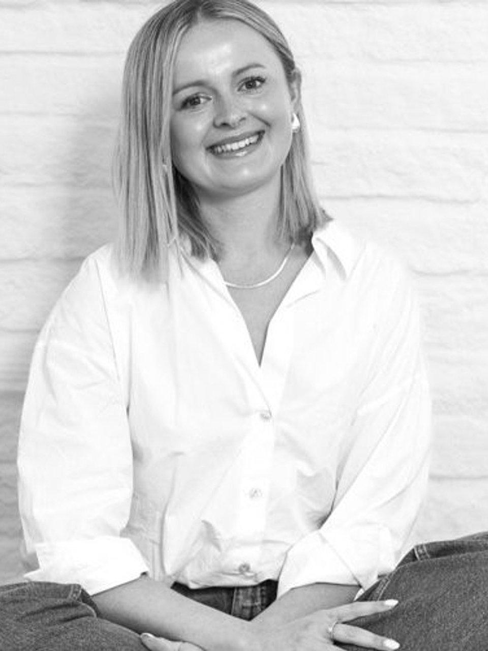 Smiling woman in a white button-down shirt and jeans seated against a white brick wall