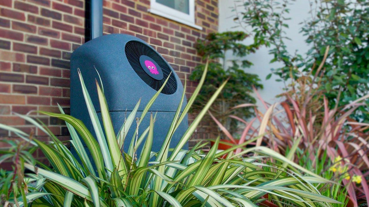 An outdoor heat pump unit beside a brick house, surrounded by garden plants.