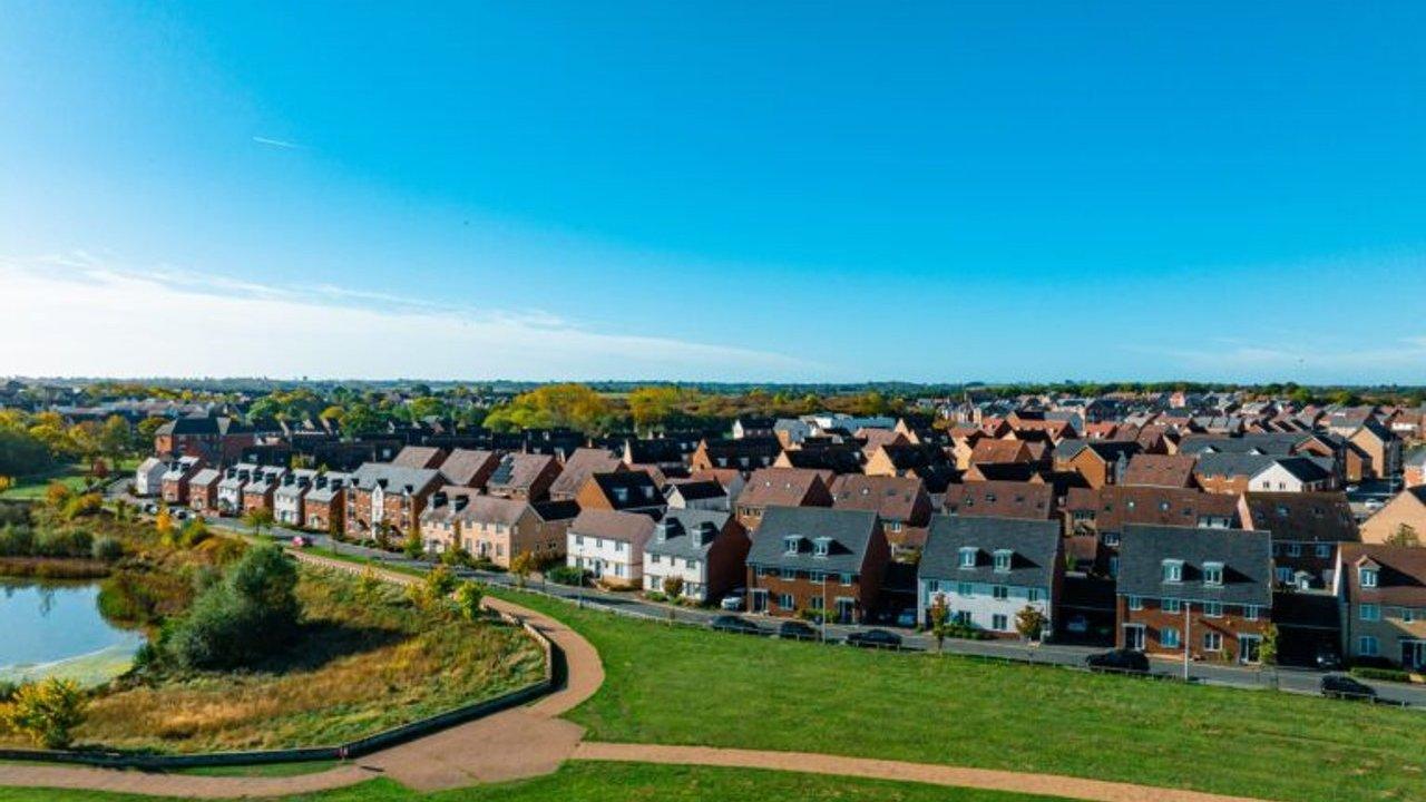 Rows of suburban houses beside a pond, green parkland and walking paths under a blue sky
