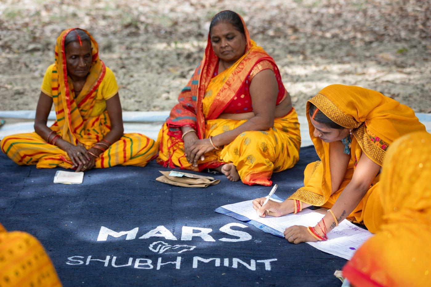 Women from a Shubh Mint self-help group sitting on a Mars Shubh Mint mat, one writing in a notebook