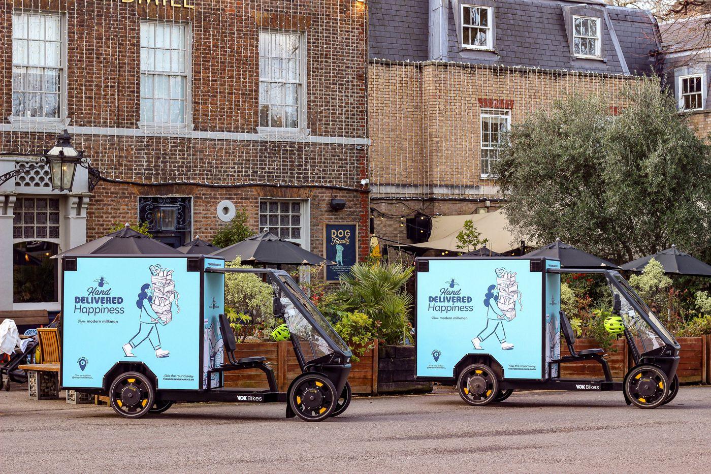 Two Modern Milkman cargo delivery bikes branded Hand Delivered Happiness, parked outside a brick pub