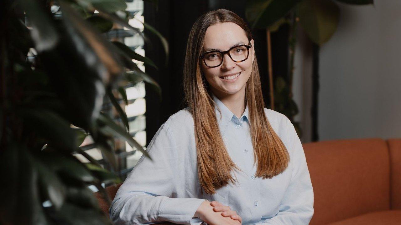 Ruta Grigaliunaite wearing glasses and a light blue shirt, seated beside leafy plants.