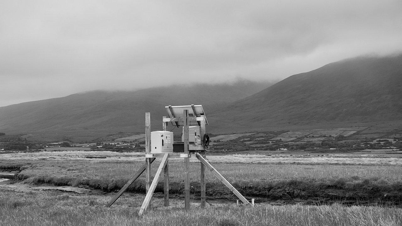 Tide-monitoring equipment on a wooden platform in a grassy marsh beneath misty mountains
