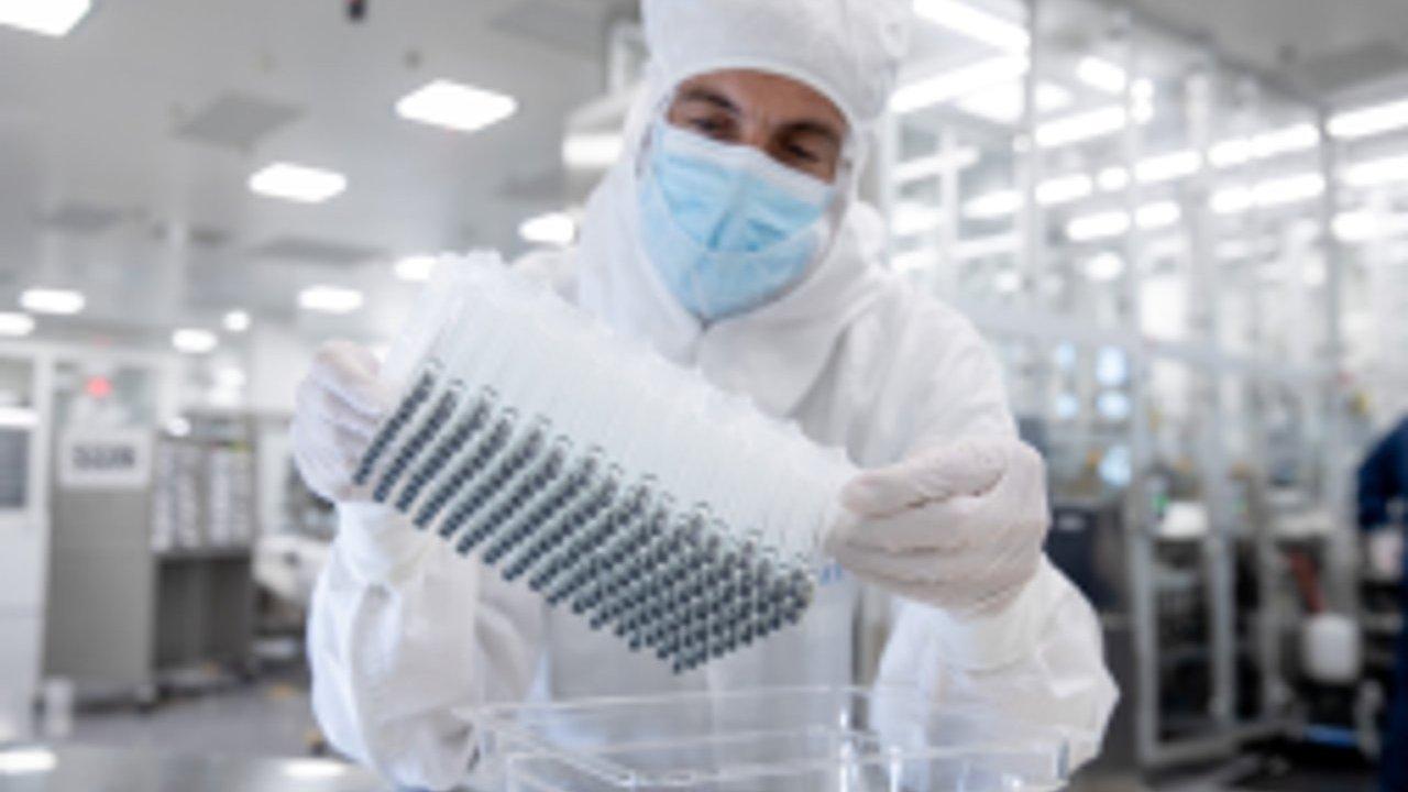 A cleanroom worker in protective gear holds a tray of small electronic components.